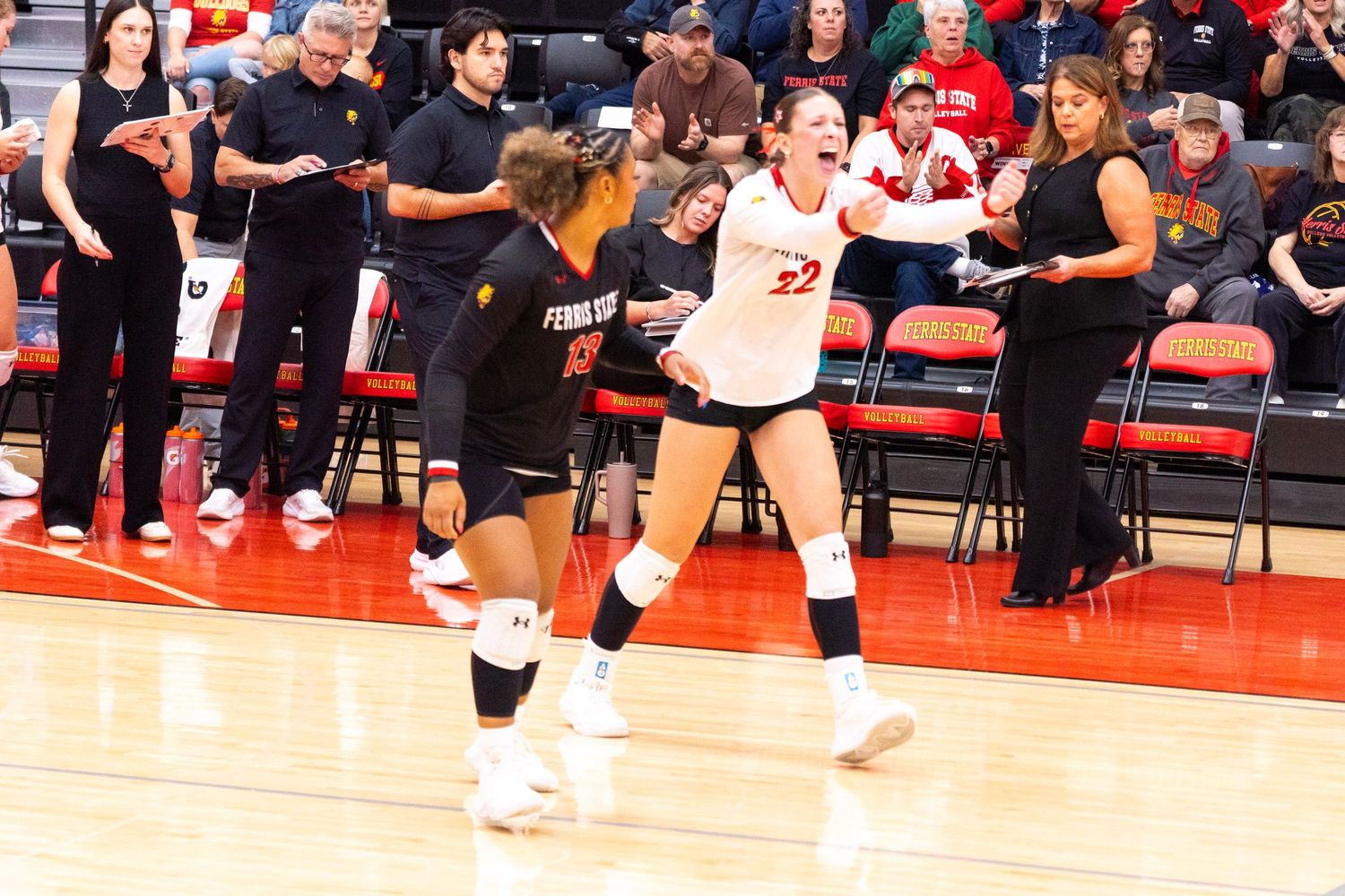 Two volleyball players in white and black uniforms celebrate a point on the court while coaches and spectators watch from the sidelines.