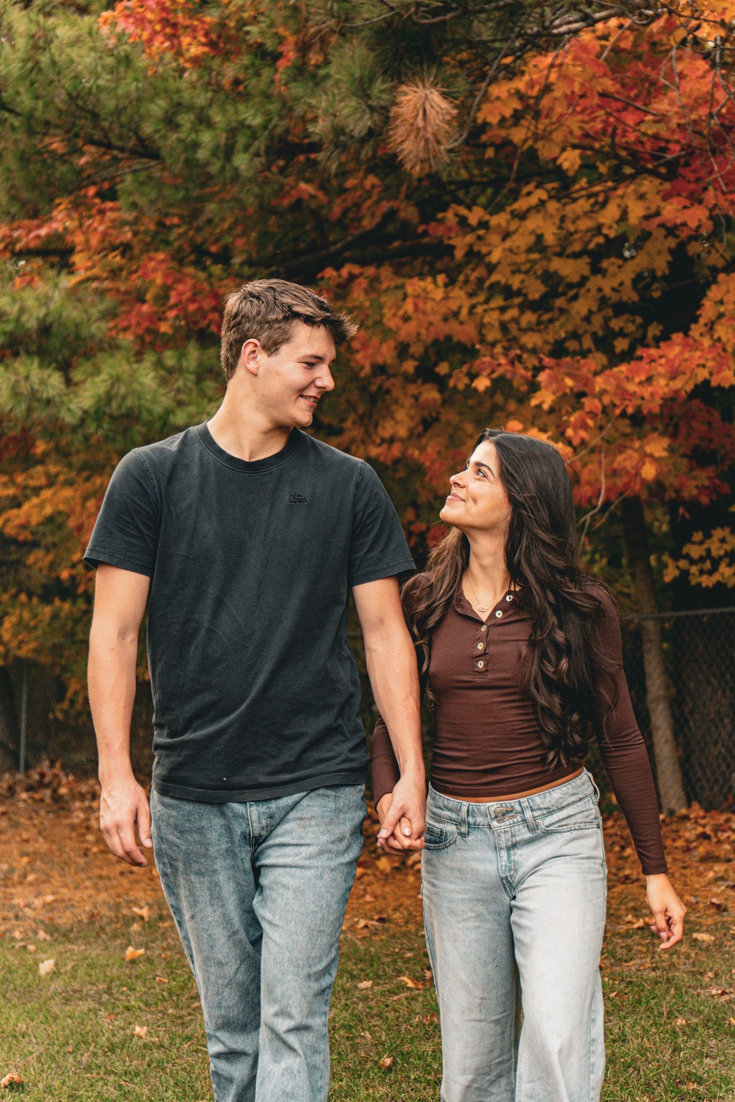 Smiling young couple holding hands and walking together on a grassy path beneath colorful autumn trees with red and orange fall foliage.