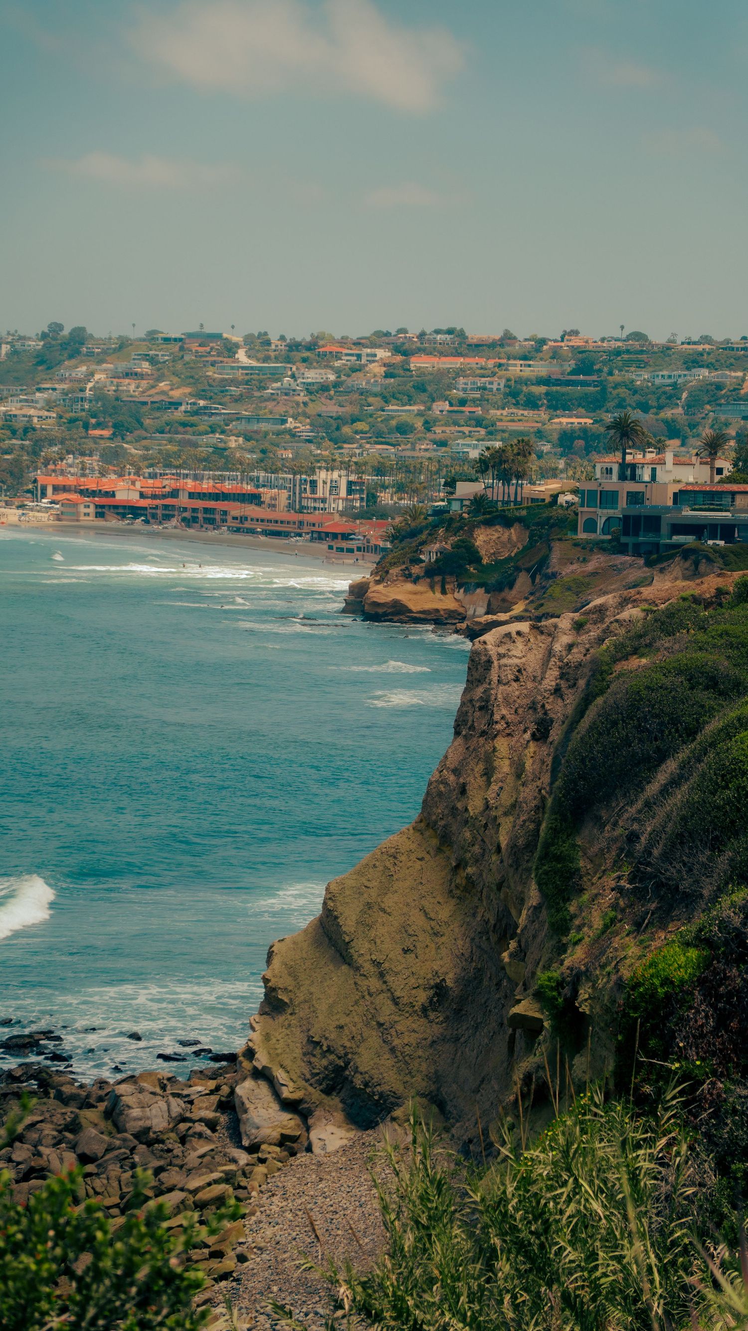Coastal homes perch atop dramatic cliffs overlooking a curved beach and blue ocean.