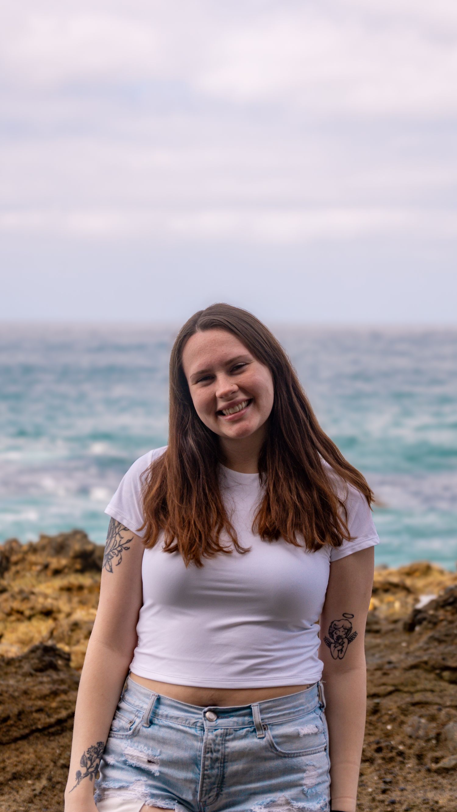 Casual beachside portrait with tattoos visible against turquoise ocean backdrop.
