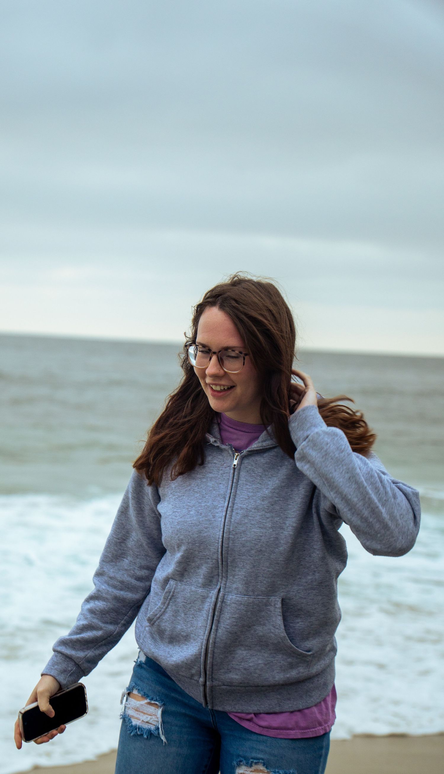 Figure wearing a gray hoodie and jeans walks along a misty beach shoreline.