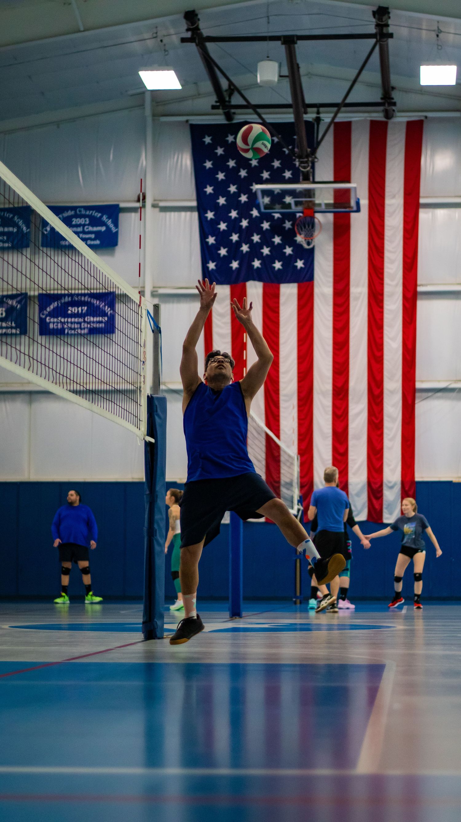 Volleyball player jumps to set ball in front of American flag during indoor game.