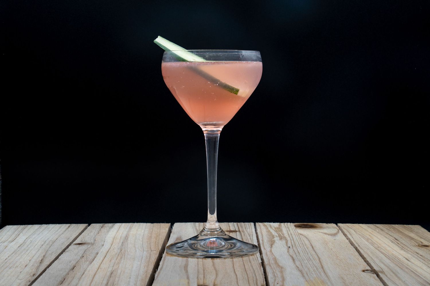 Pink cocktail in martini glass with lemongrass garnish on rustic wooden surface.