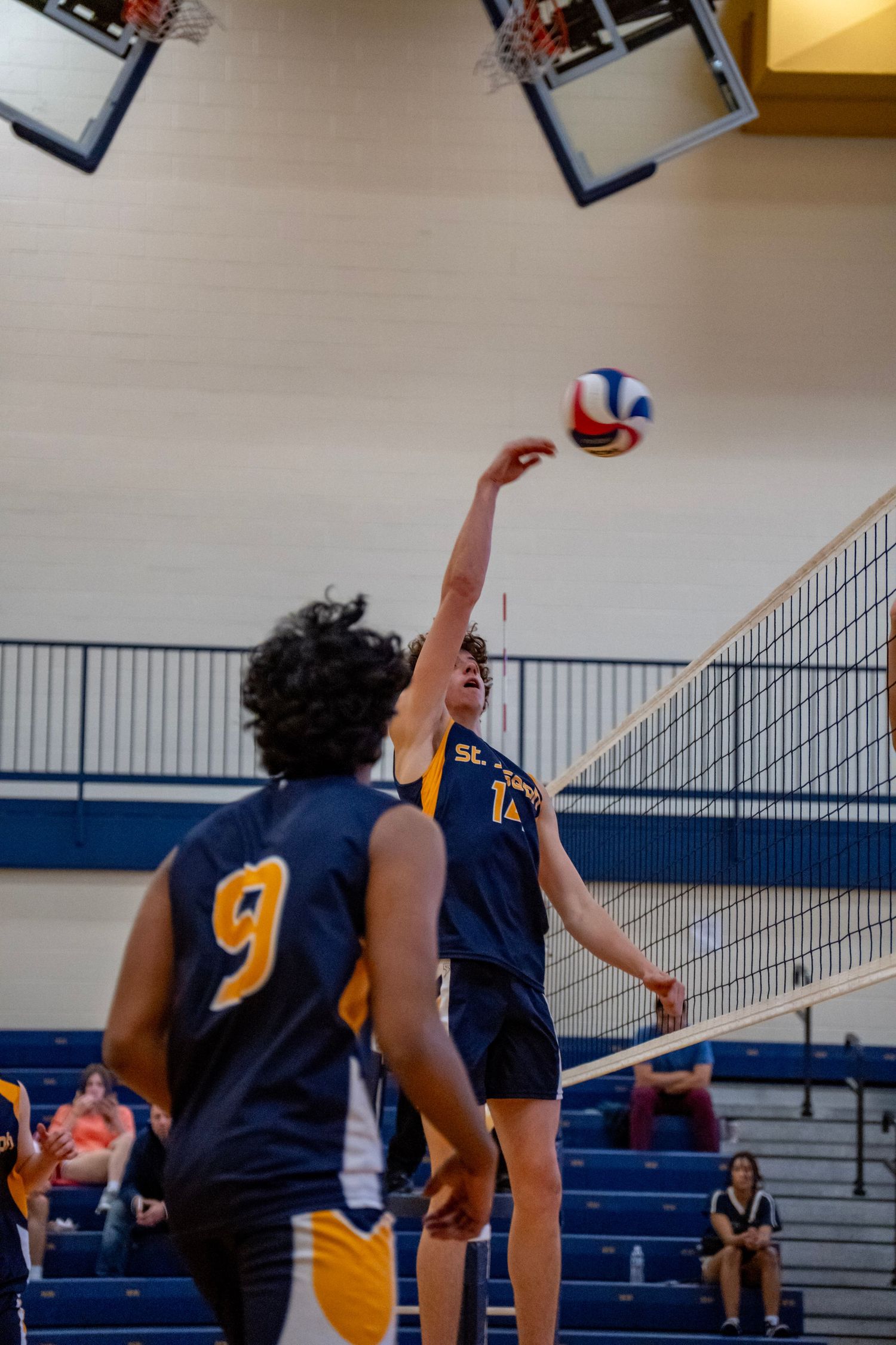 Volleyball players in navy and gold uniforms jump to block a spike during an indoor match.