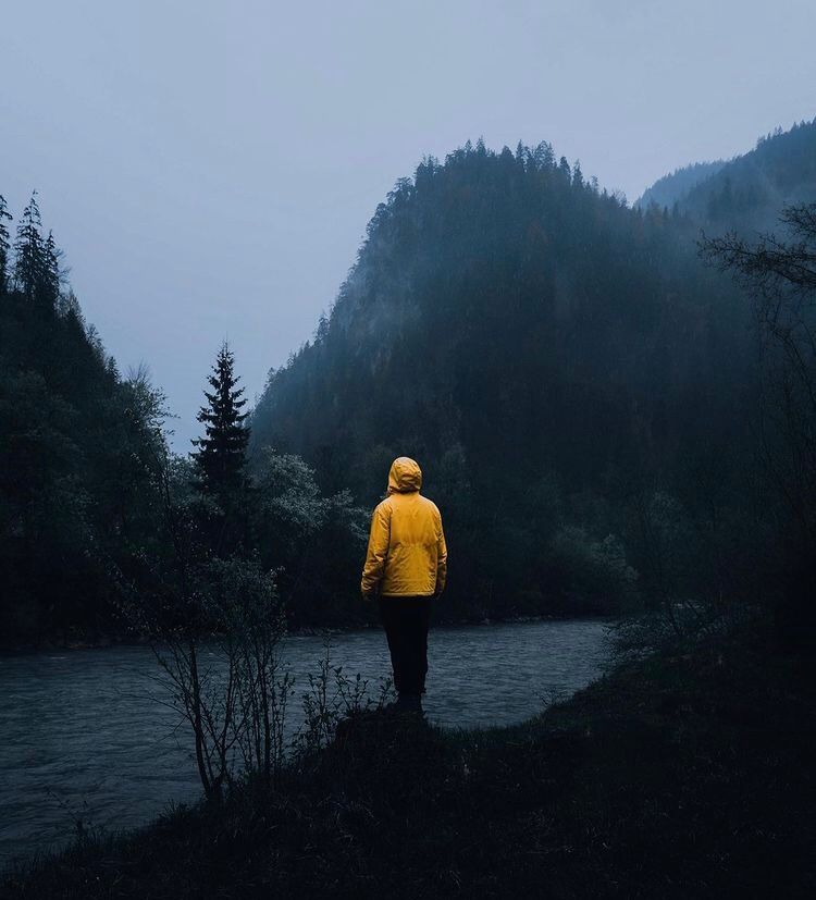 Person in bright yellow raincoat standing on cliff edge overlooking misty mountain landscape.