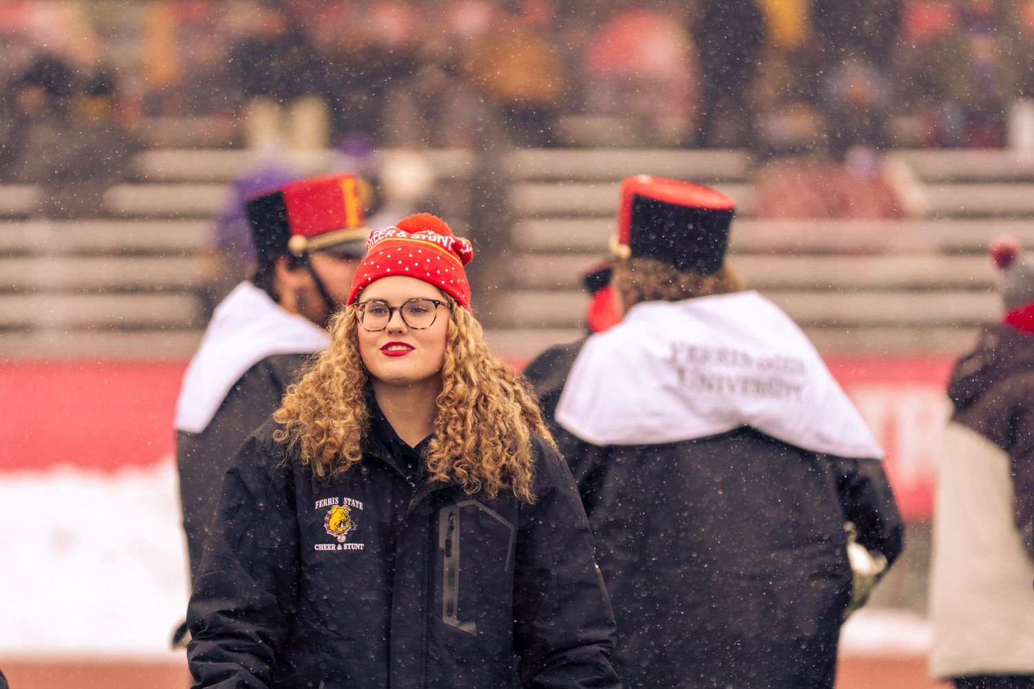 A person in a red coat and glasses stands in front of a crowd at what appears to be a sporting event.