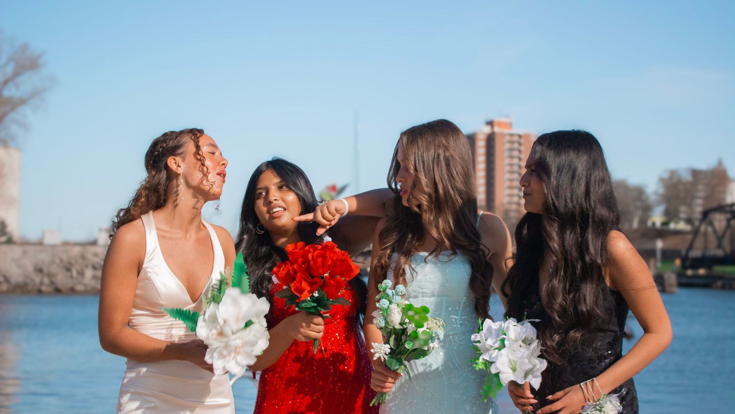 Wedding party in formal attire poses together by waterfront with city skyline visible in background.