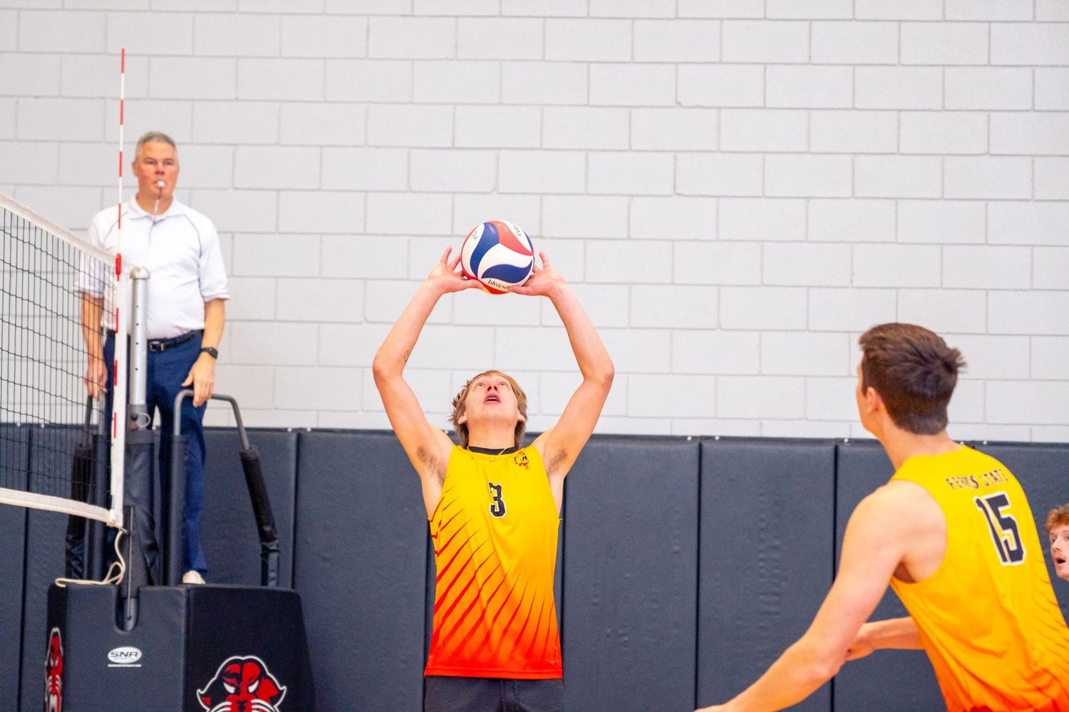 Indoor volleyball players in yellow jerseys compete at the net while a referee observes from the sideline.
