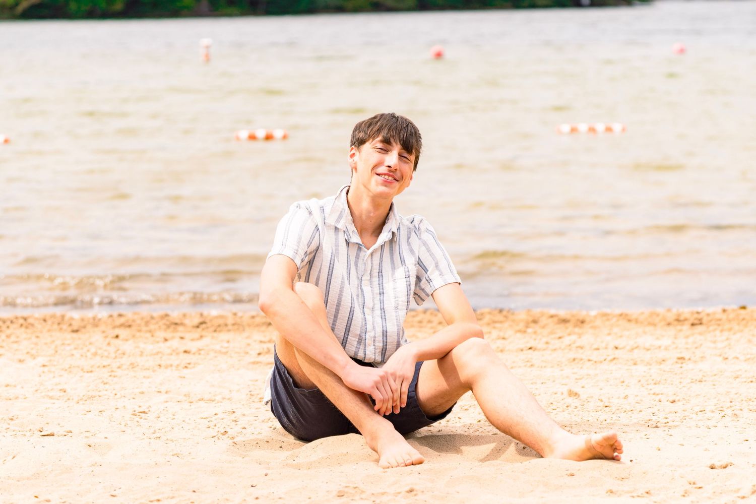 Smiling young man in a striped short-sleeve shirt and dark shorts sits barefoot on a sandy beach with calm lake water and buoys behind him.