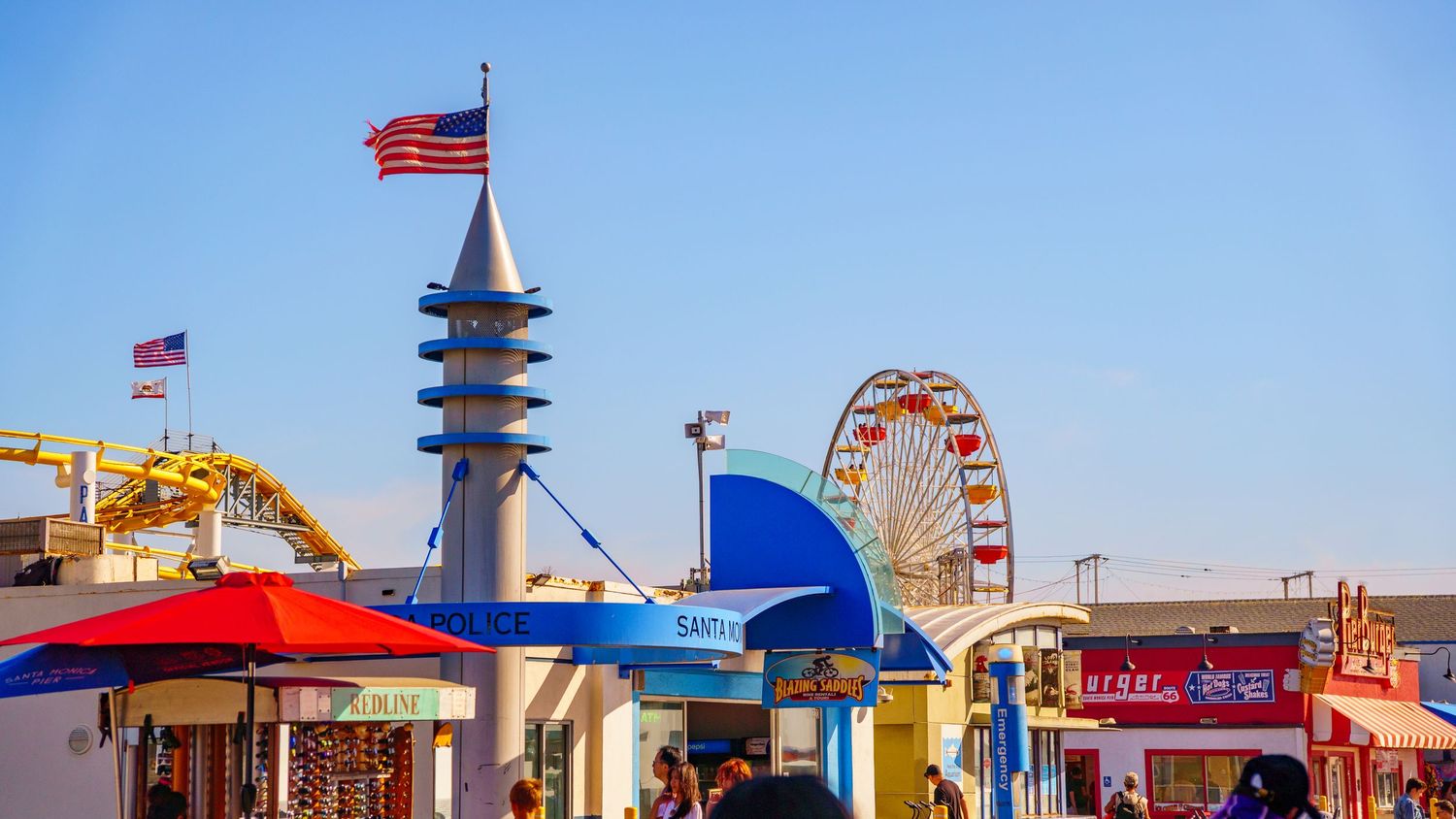 American flag flies over retro-styled buildings and Ferris wheel at seaside amusement park.