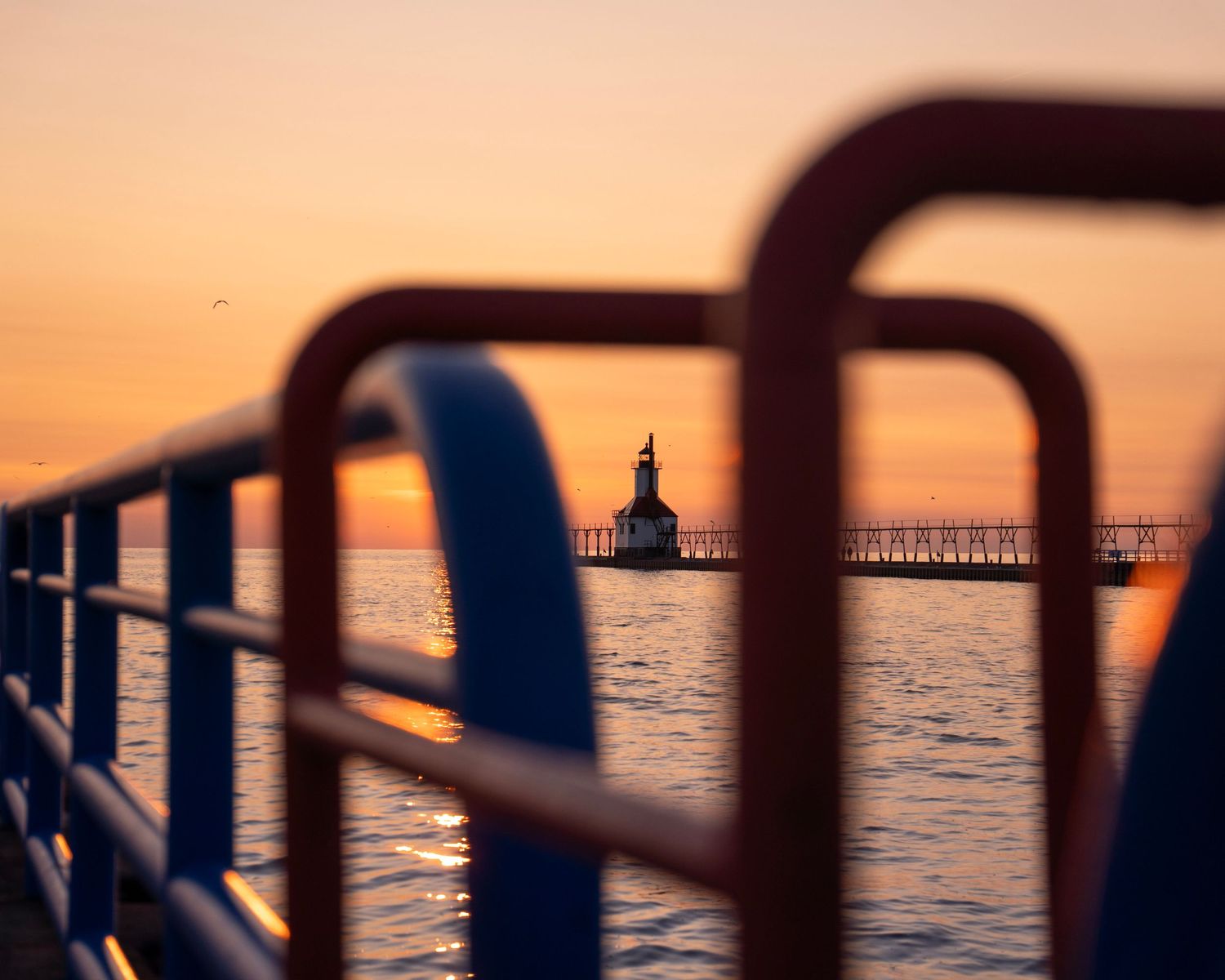 Blue railing frames lighthouse and pier during golden hour on Lake Michigan.