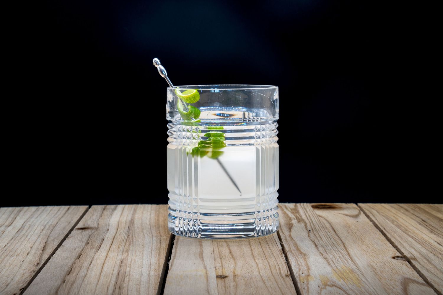 Clear cocktail with fresh herbs in textured crystal glass on rustic wooden surface.