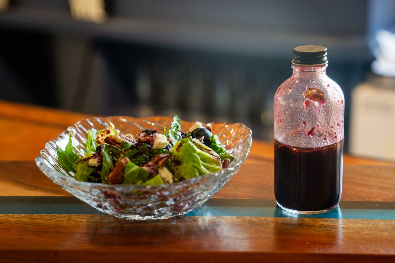 Fresh garden salad in crystal bowl with dark liquid in glass bottle on wooden table.
