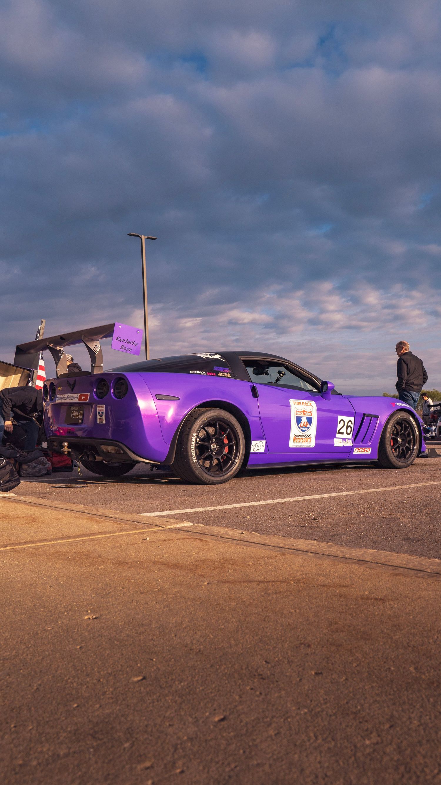 Purple Corvette race car with black wheels parked at a motorsport event under cloudy skies.