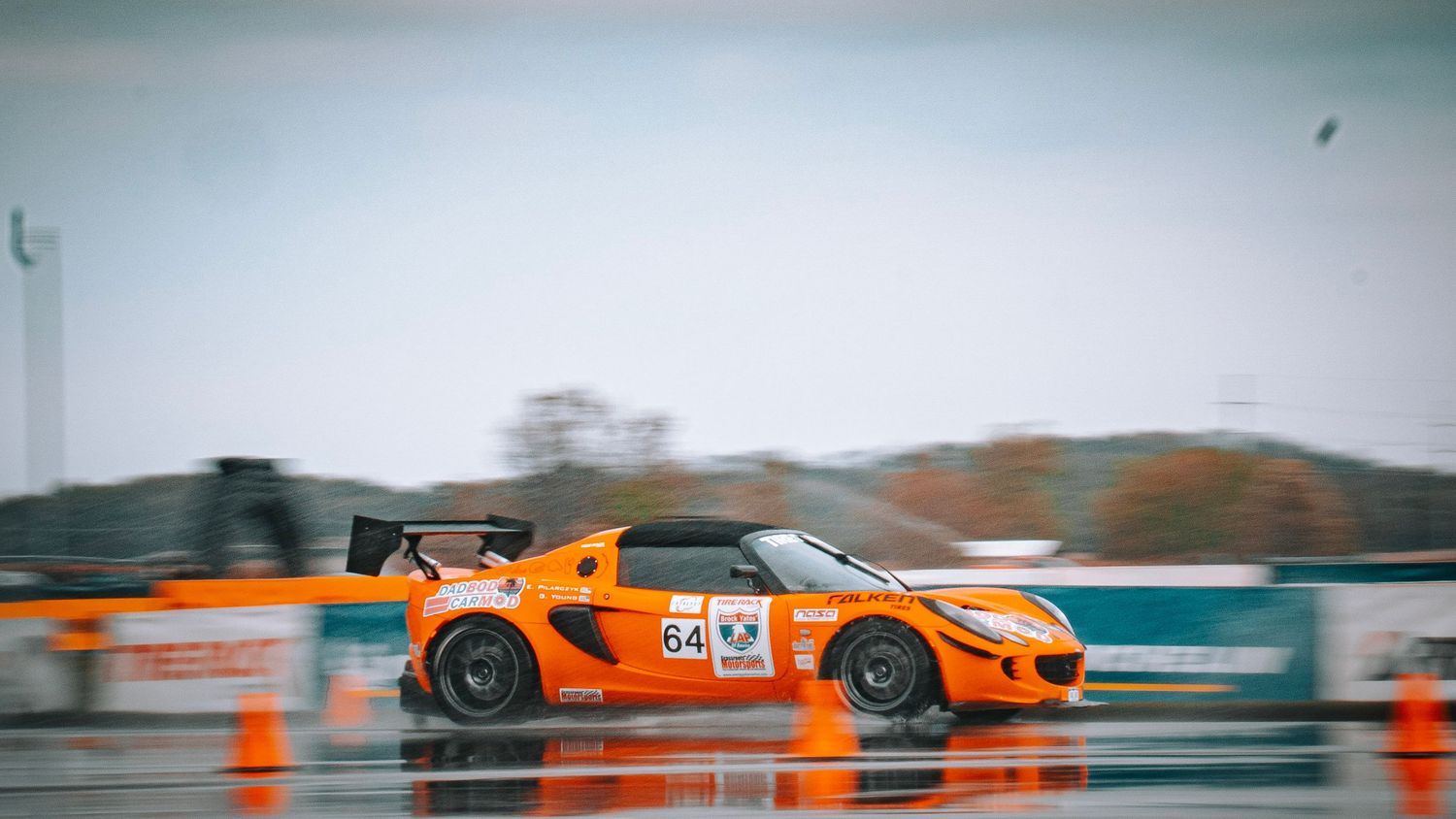 Orange racing car navigating through wet track with cones during rainy conditions.