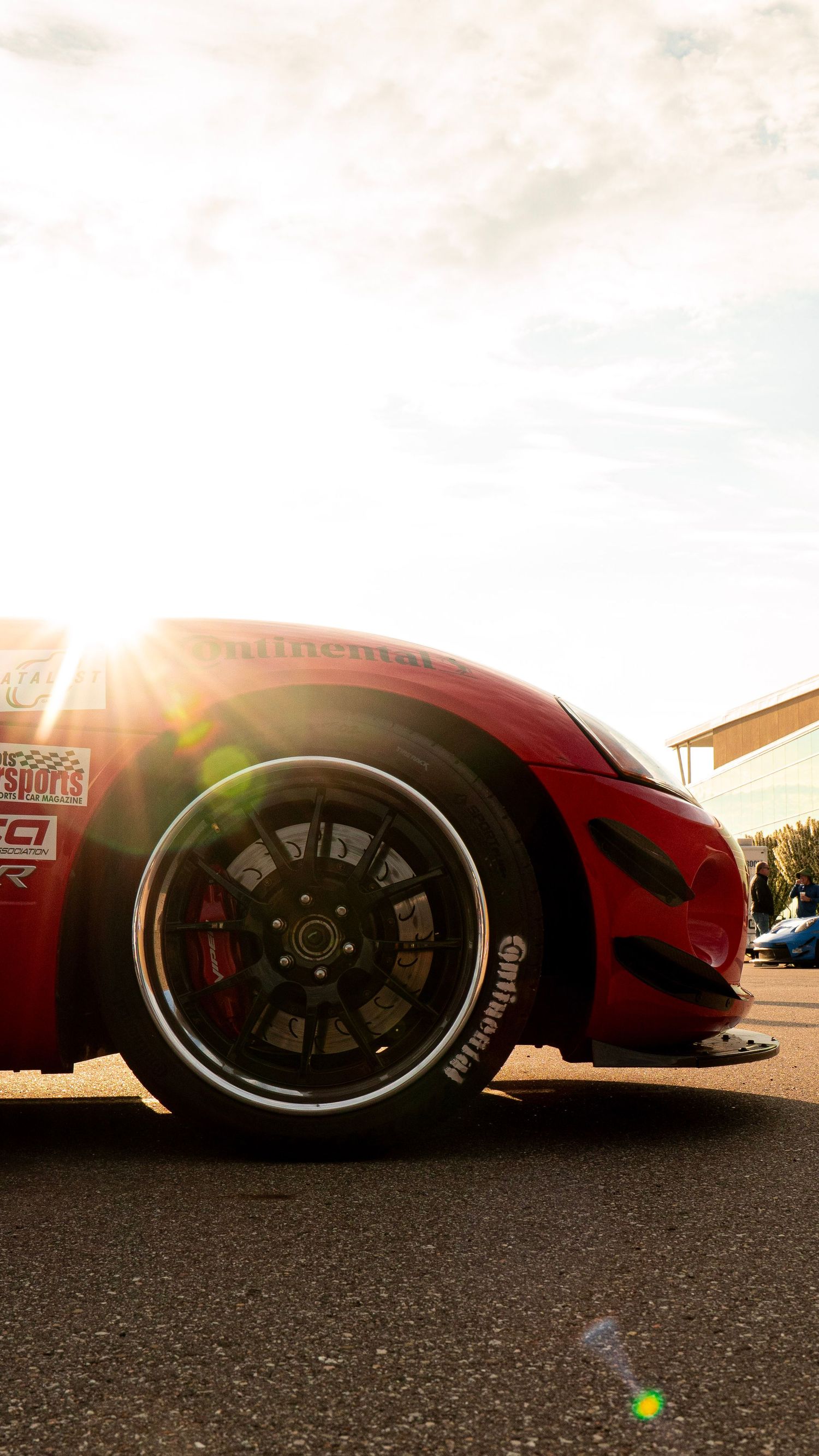 A red sports car wheel with glowing sunset light creating lens flare against dark asphalt.