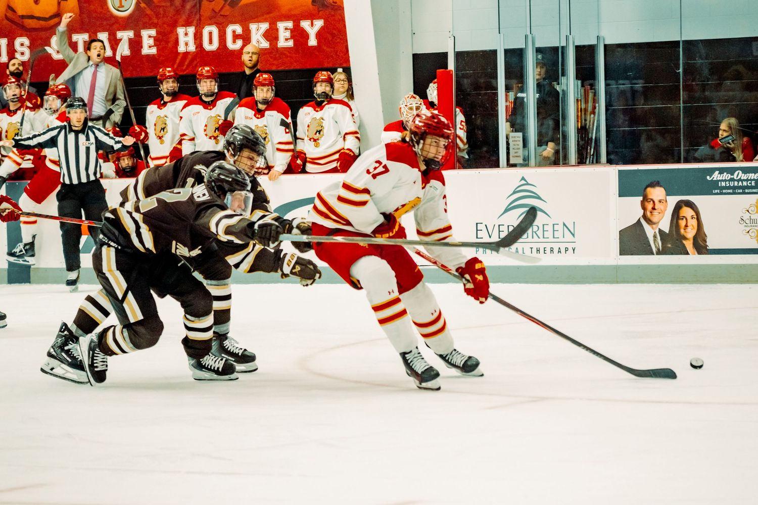 A college hockey player in a red and white jersey chases the puck on the ice while a defender in a dark uniform pursues him.