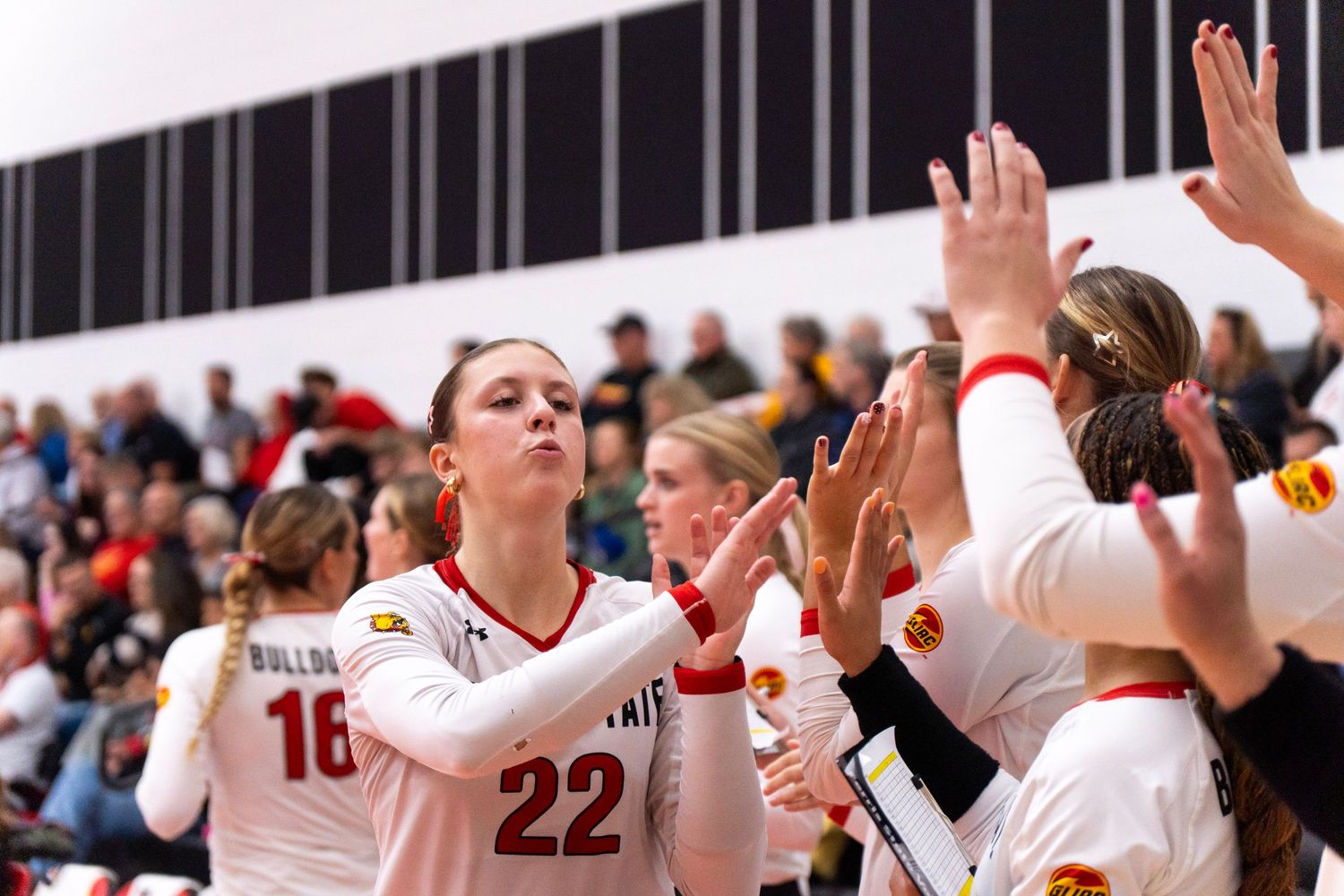 Volleyball player in white number 22 jersey exhales while teammates reach out to high-five on the court as fans watch from the stands.