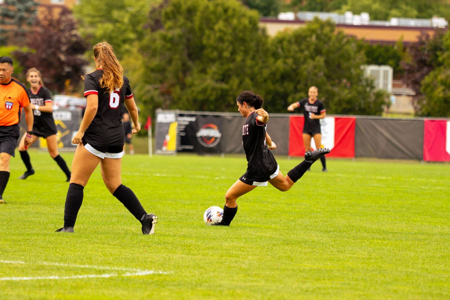 Female soccer player in black kicks the ball on a green field as teammates and an opponent look on during a college game.
