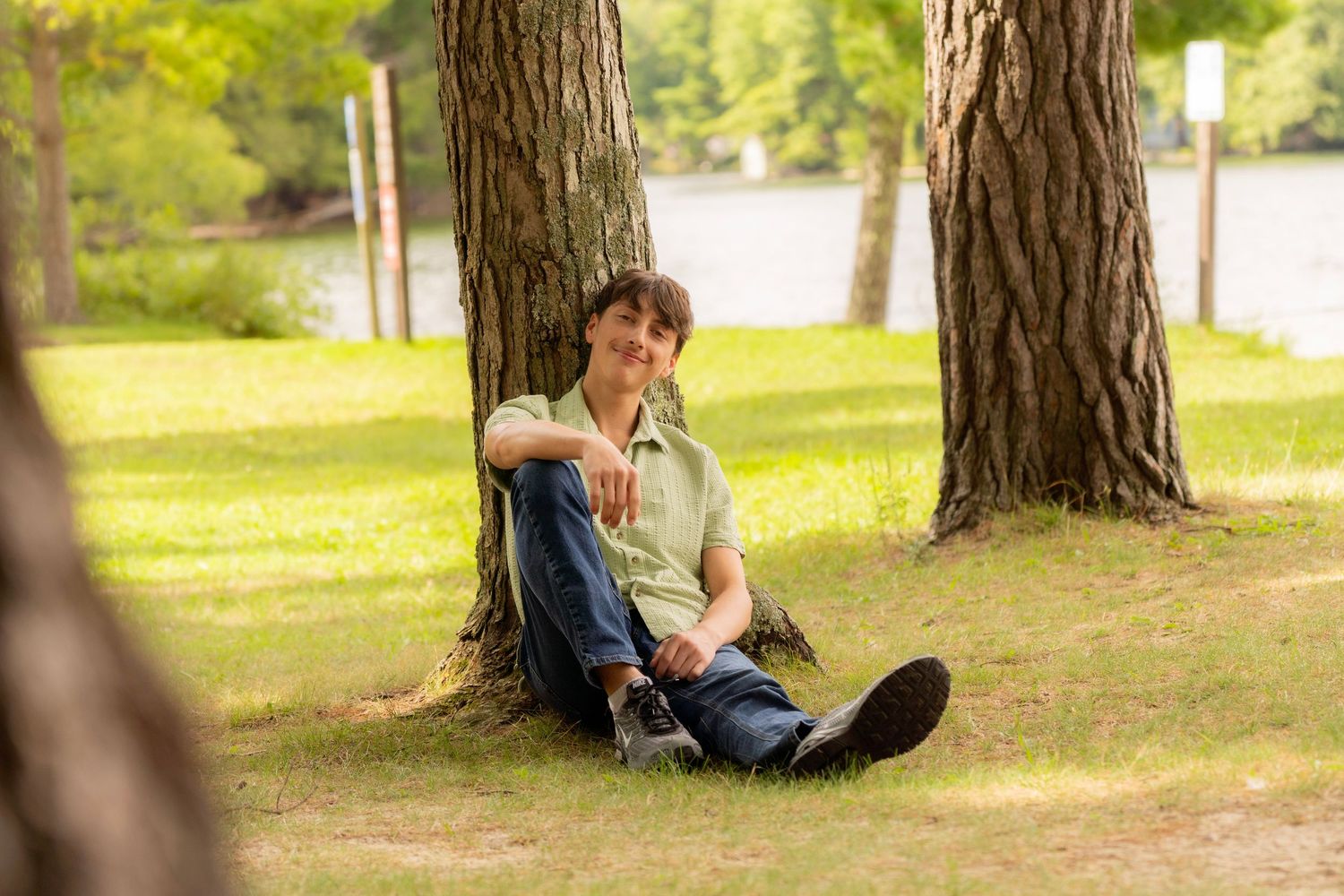 Smiling young man in a green shirt and jeans sits against a large pine tree on a sunny grassy lawn near a calm lake.