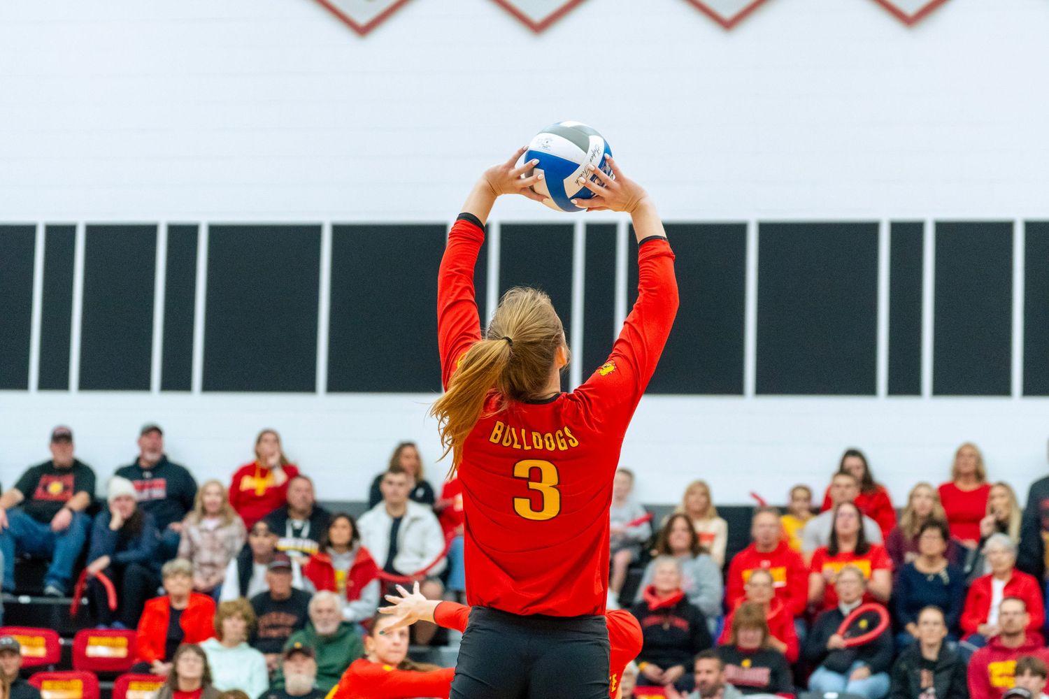 Volleyball players in red uniforms jump to block during an intense match against opponents.