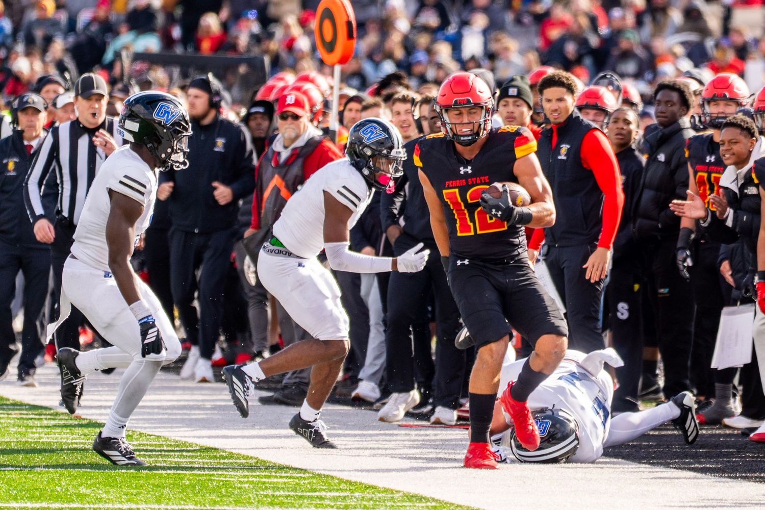 Football players dive for the ball during an intense play along the sideline with spectators watching closely.