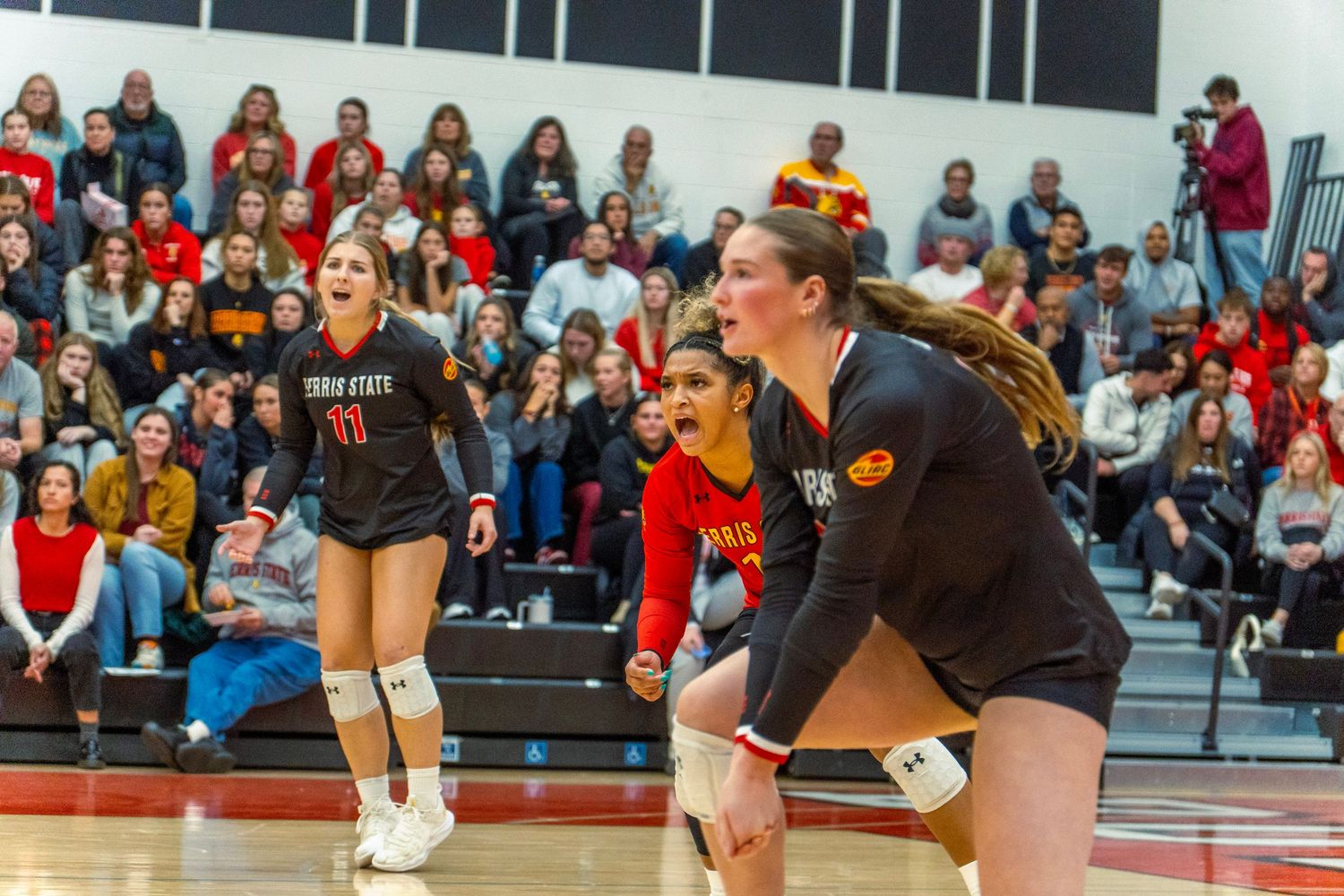 High school volleyball players compete at the net during an intense match with spectators watching from the bleachers.