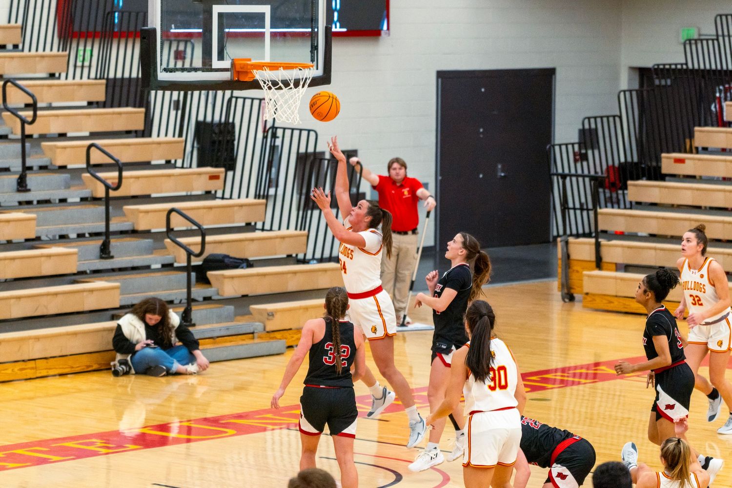 Basketball players compete during an indoor game with a player shooting the ball while others watch from wooden bleachers.