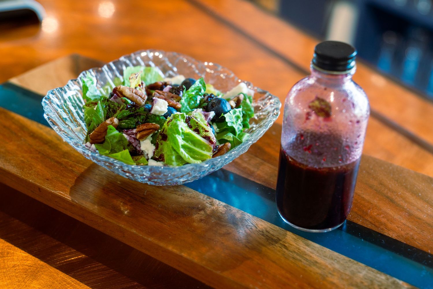 Mixed green salad in crystal bowl beside bottle of dark dressing on wooden surface.