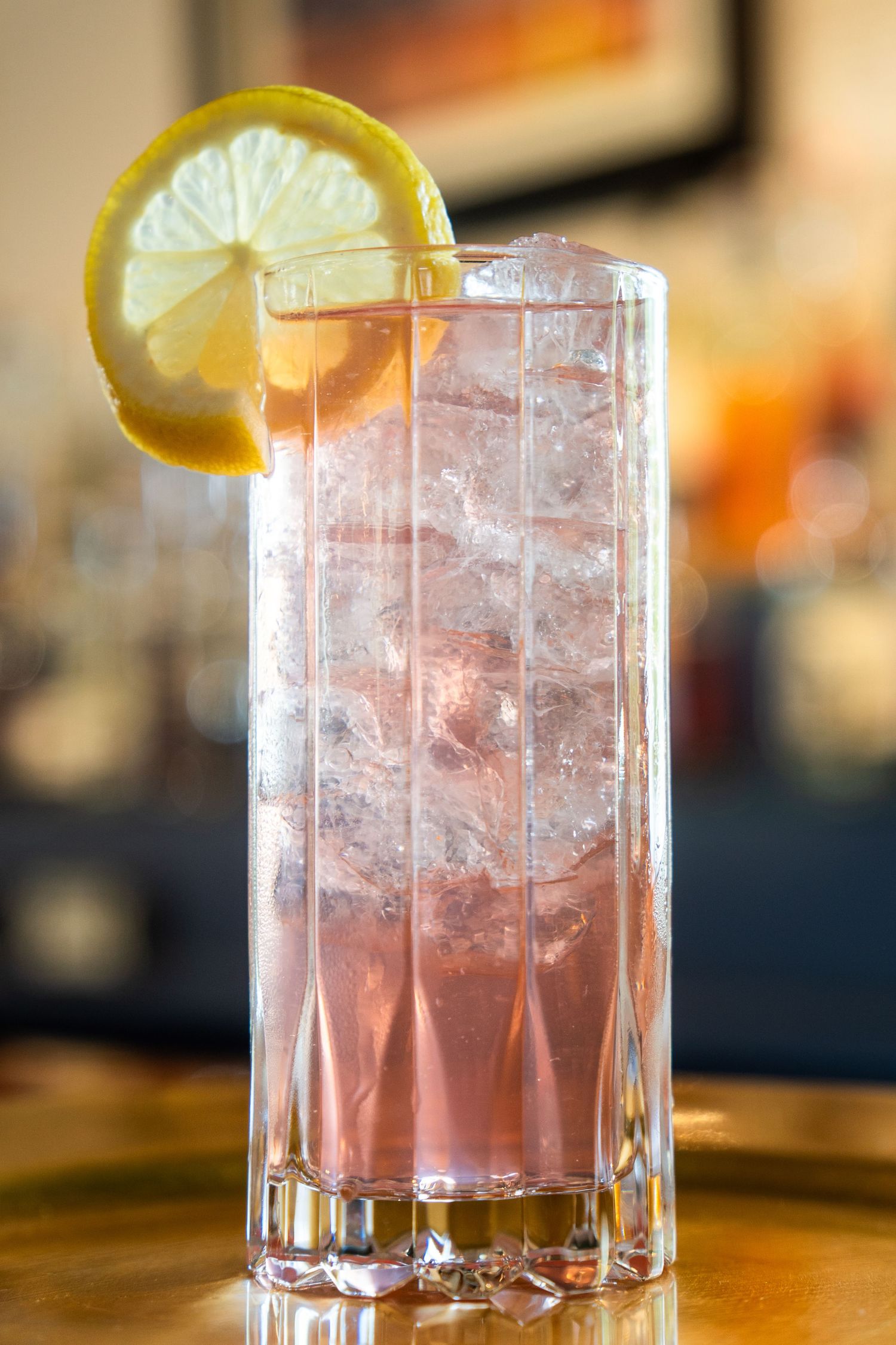 Pink cocktail with ice and lemon garnish in a tall crystal glass.