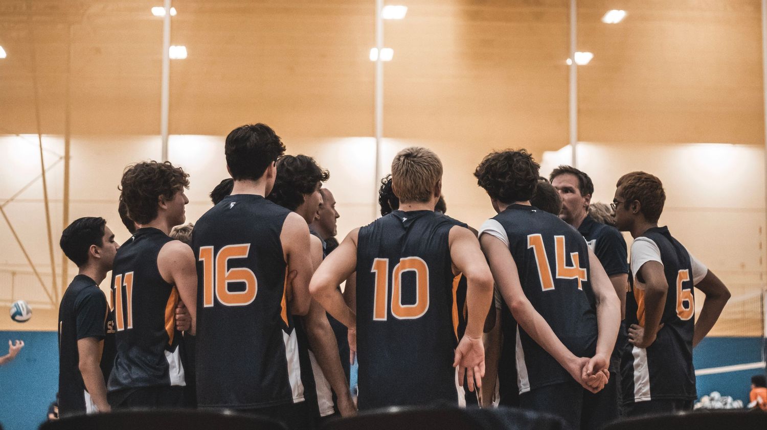 Team huddles together wearing matching black and orange jerseys with numbers.