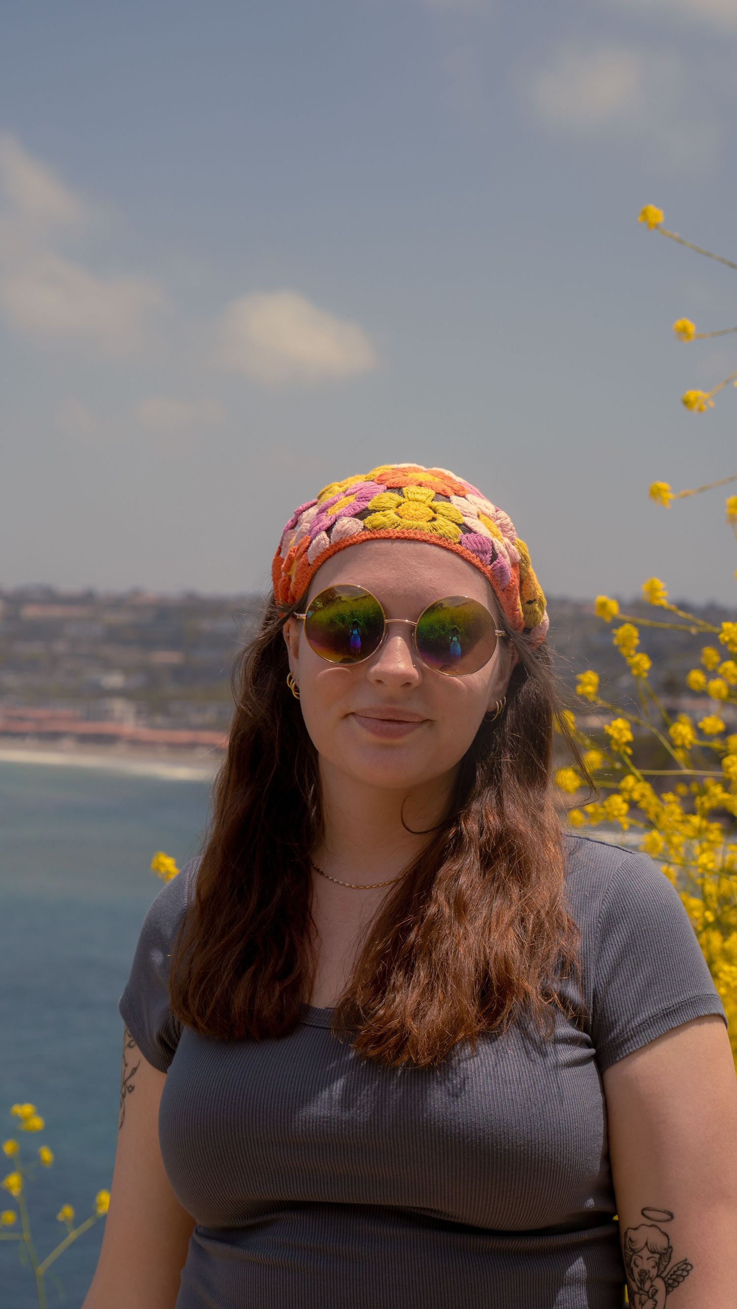 Person in colorful headscarf and sunglasses smiles with ocean and yellow flowers in background.
