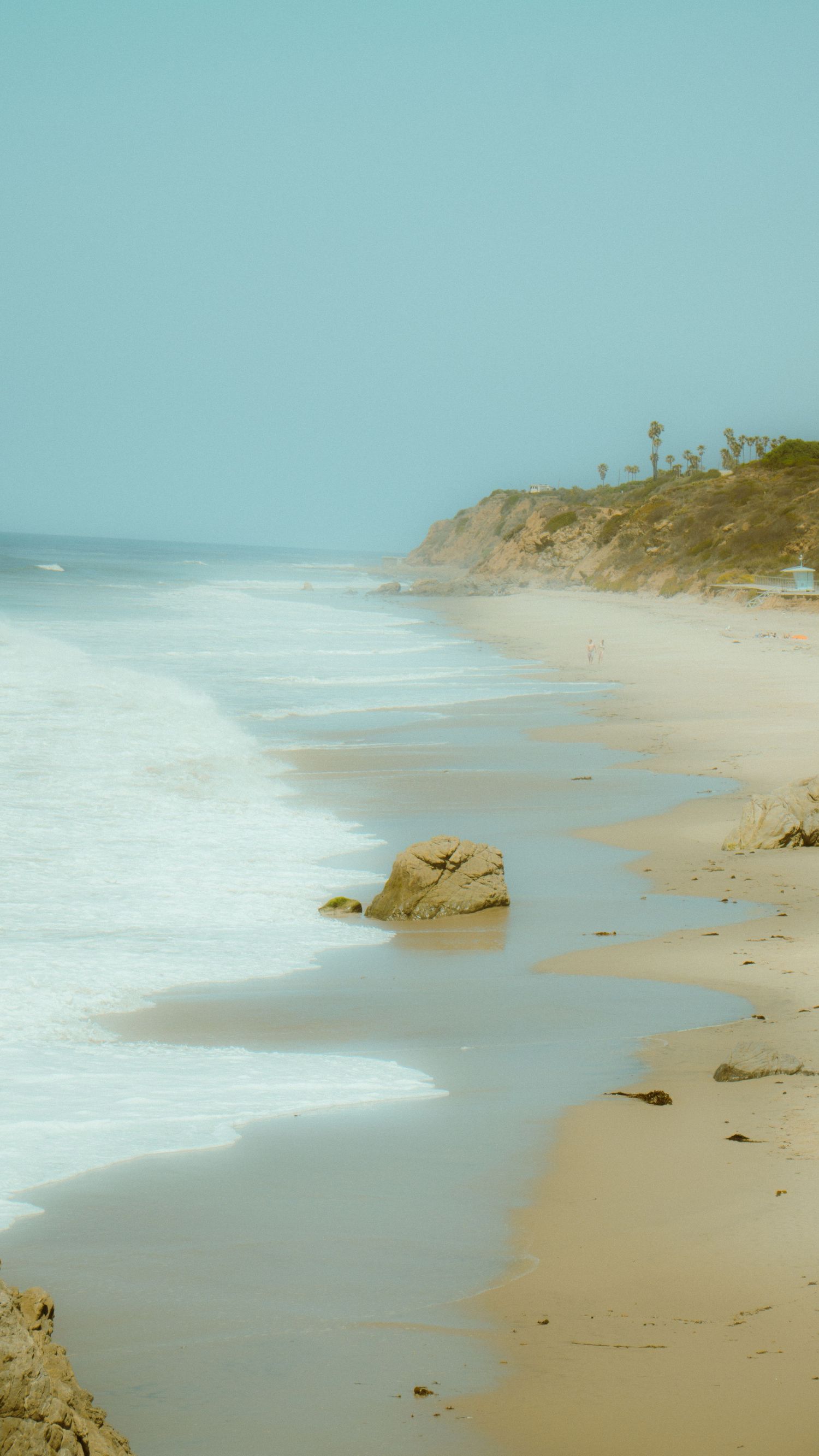 Pristine sandy beach stretching along turquoise waters with rocky outcrops in the distance.