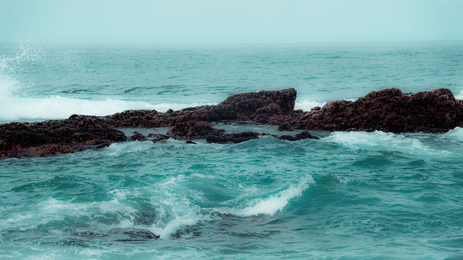 Turbulent turquoise waters crash over dark rocky outcrops creating dramatic ocean spray.