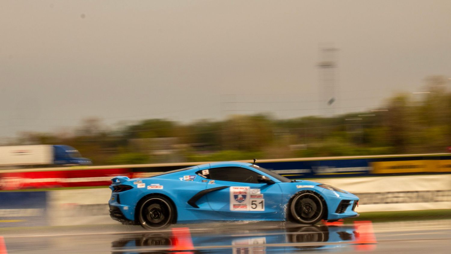 Blue race car speeds past orange barriers on rainy race track.