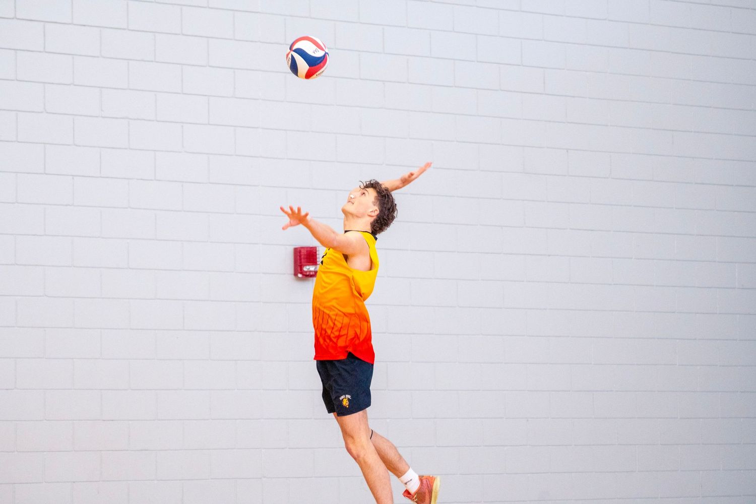 A volleyball player in an orange jersey jumps high to hit the ball against a white wall background.