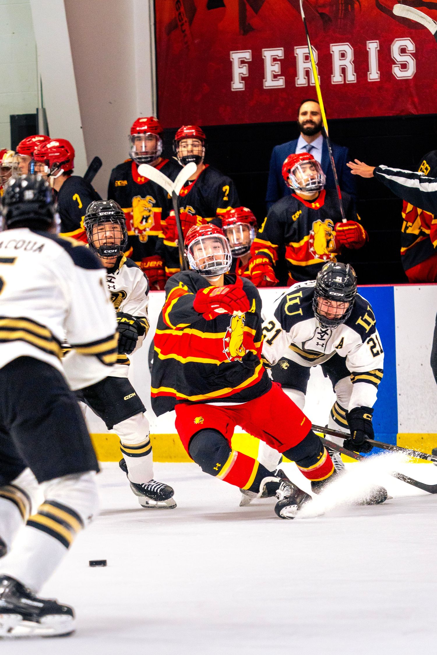 Ice hockey player in a red and gold jersey slides across the ice as opponents in white and a bench of teammates watch the game action.