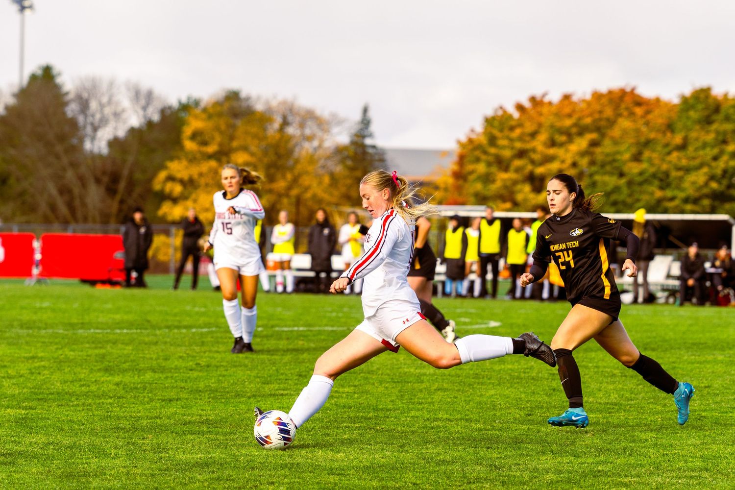 Female soccer player in a white uniform kicks the ball on a grass field as a opponent in black and gold chases during a fall match.