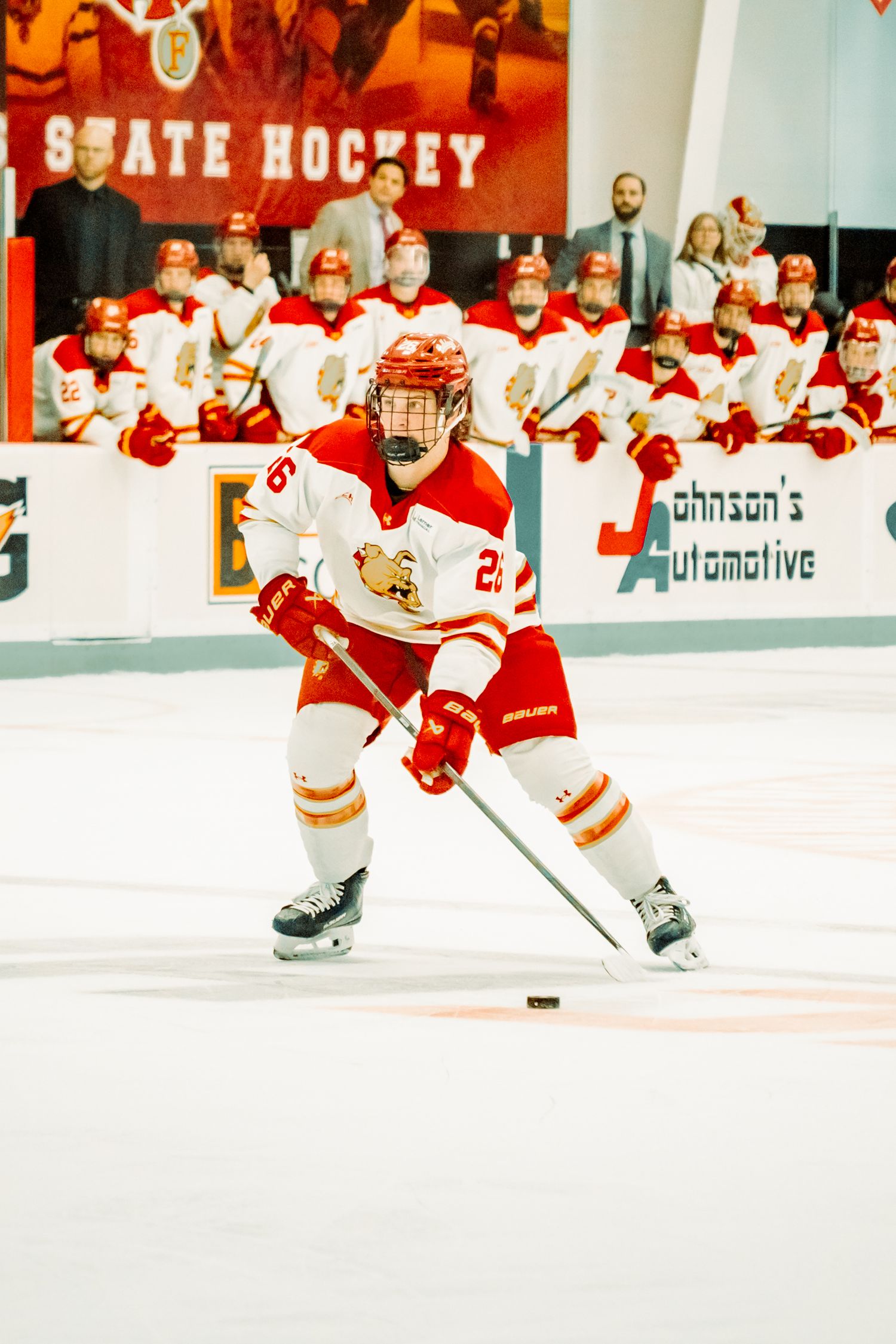 Ice hockey player in a white and red jersey skates with the puck while teammates and fans watch from the bench at a state hockey arena.