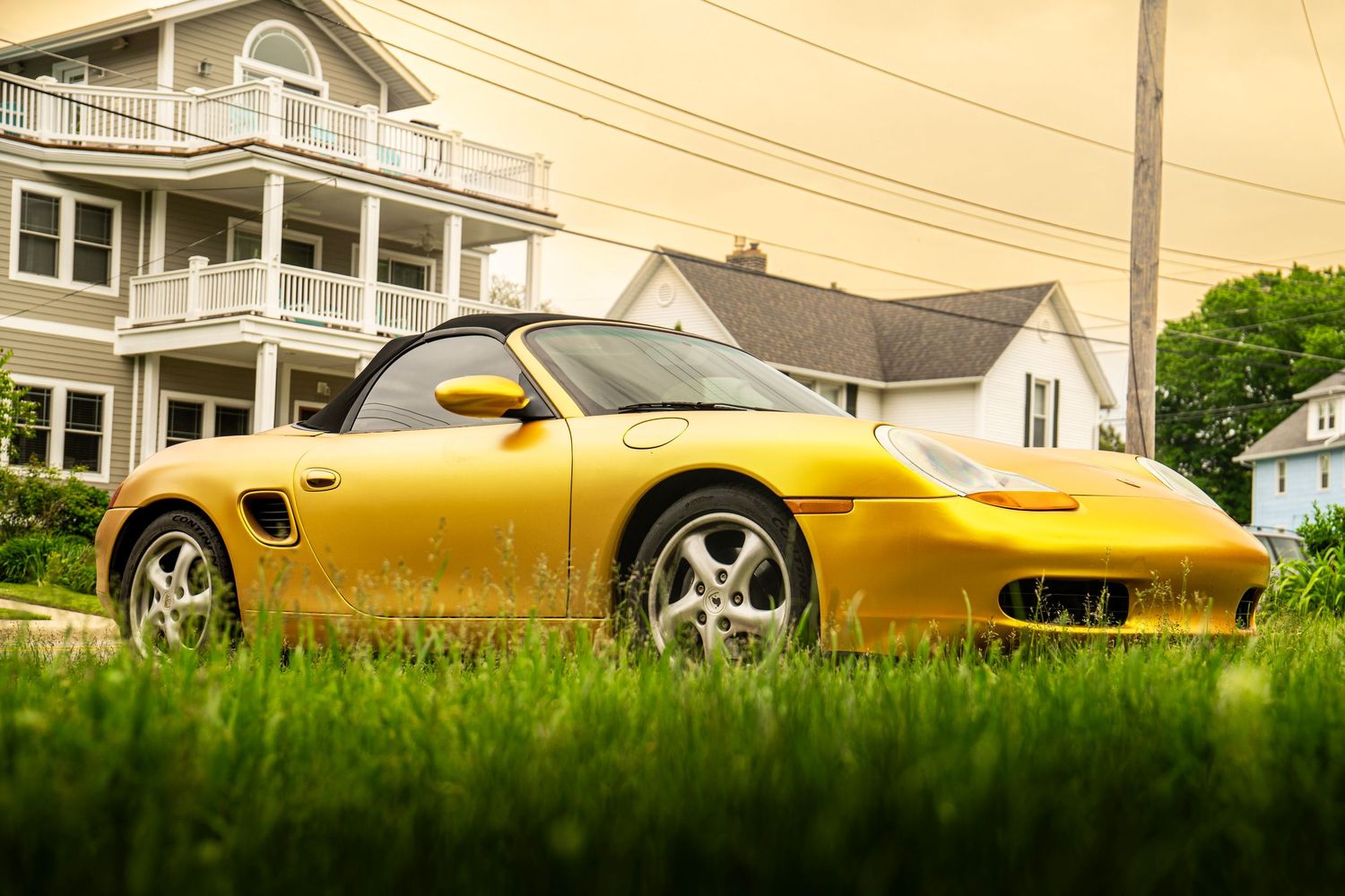 Yellow Porsche Boxster sports car parked on grass with coastal houses in background.
