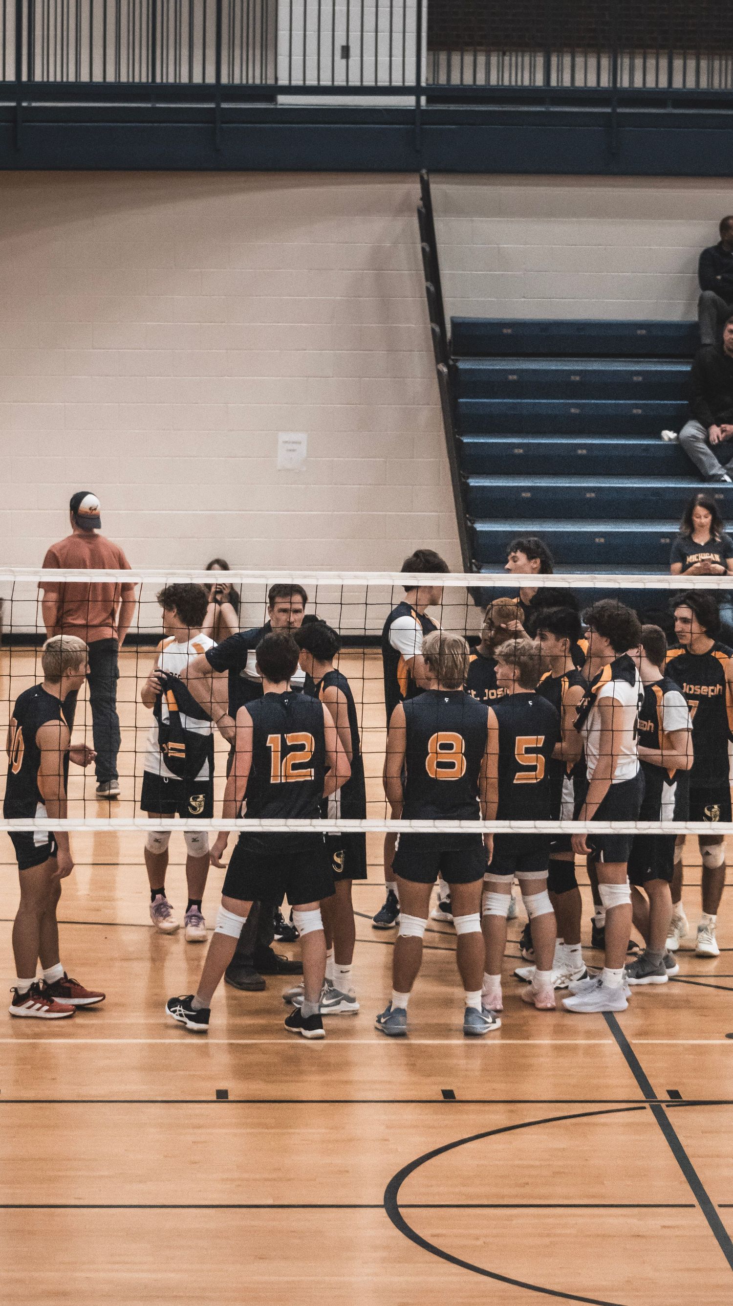 Volleyball team huddles around the net during a match, players wearing black jerseys with numbers.
