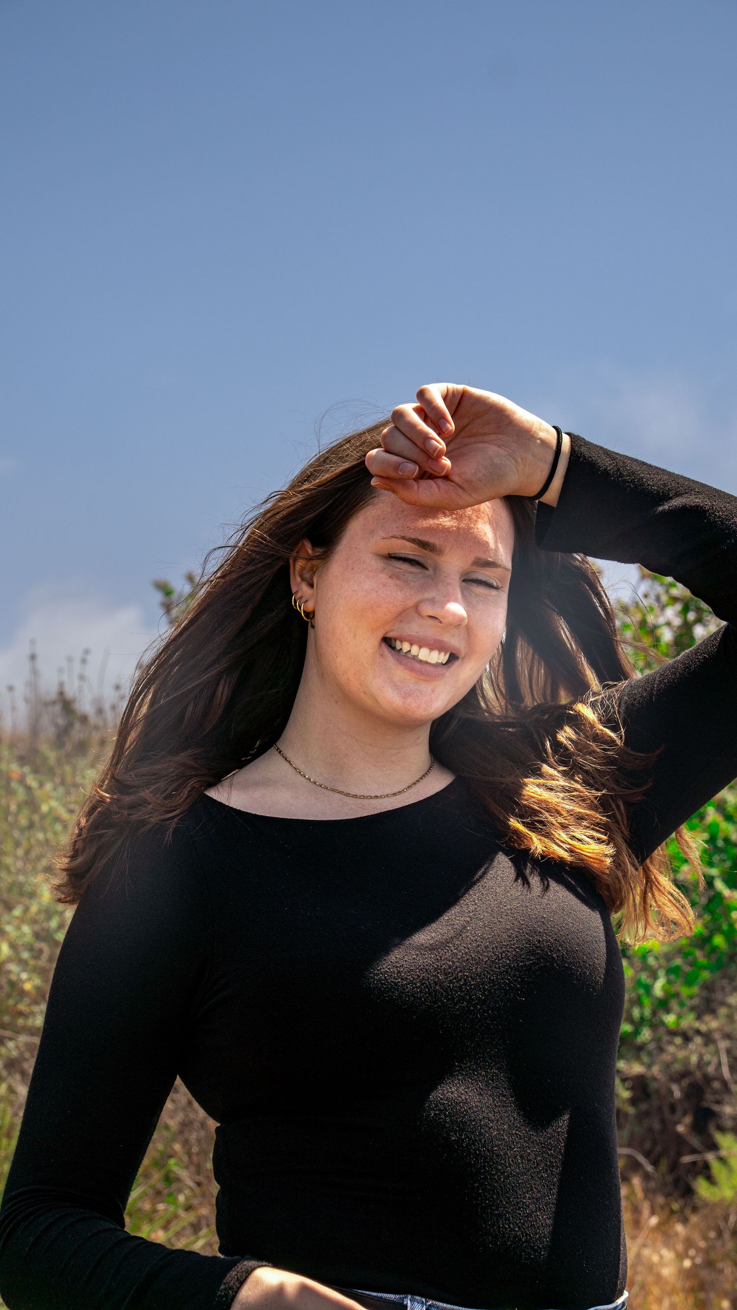 Person in black top laughing while shielding eyes from sun against bright blue sky.
