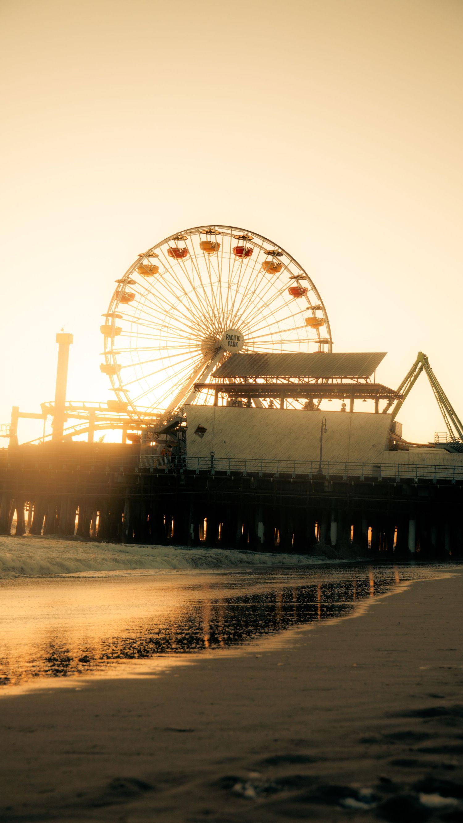 Iconic Ferris wheel and pier structures casting reflections on wet sand during sunset.