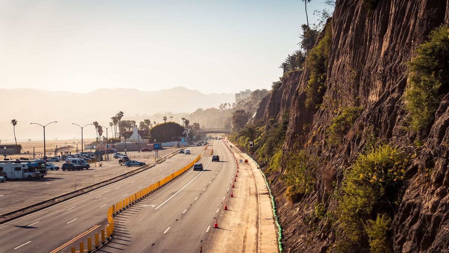 Multi-lane highway runs parallel to steep rocky cliffs with scattered vehicles.