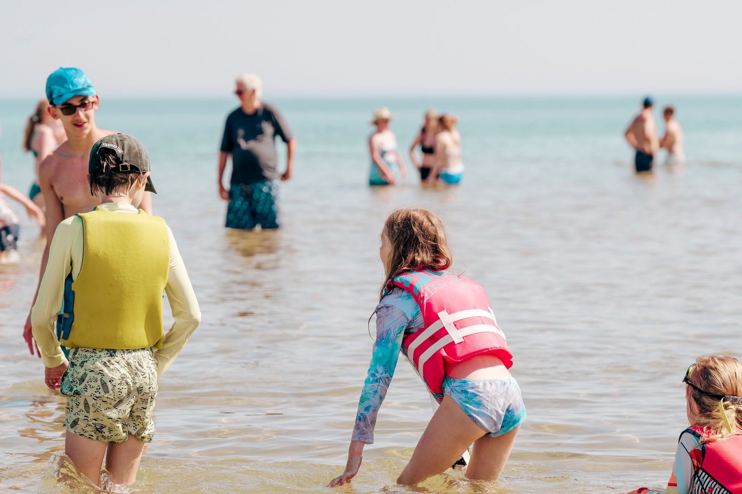 Several beachgoers wade in calm shallow ocean water on a sunny summer day.