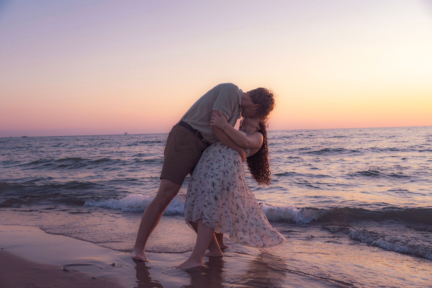 A romantic couple embraces on the beach at sunset with ocean waves gently rolling in the background.