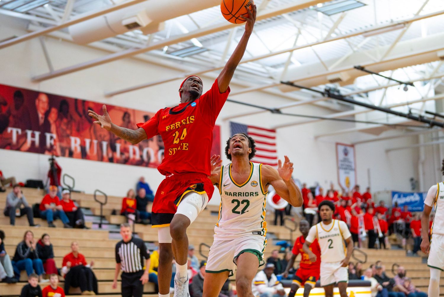 Basketball players leap high during an intense game in a gymnasium with American flags and spectators visible.