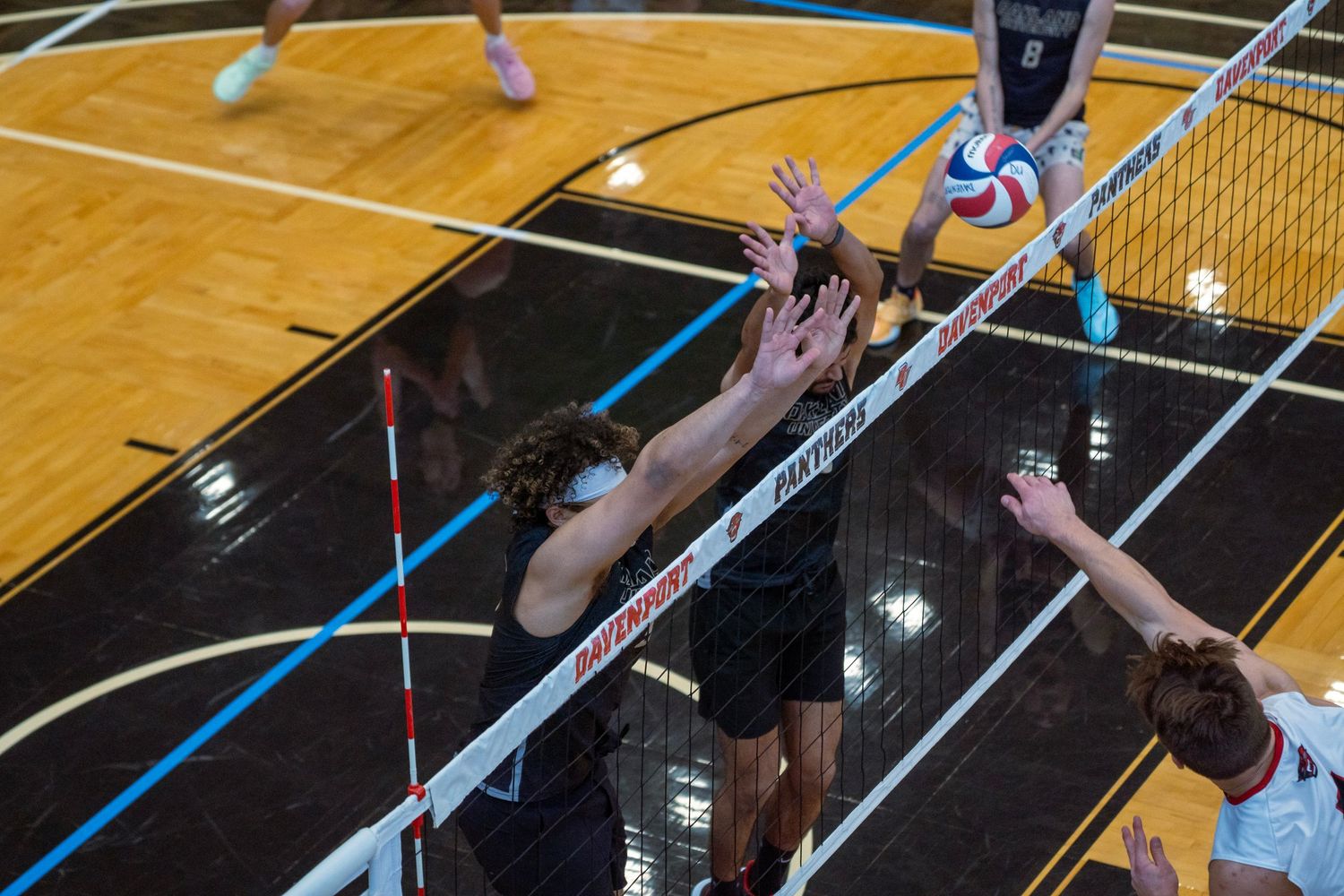 Volleyball players leap at the net during an intense match on a wooden court.