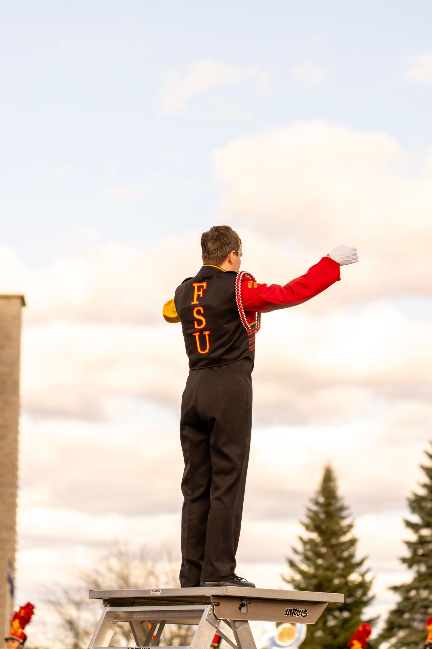 Three people in black overalls and red sleeves stand on elevated platforms against a cloudy sky at sunset.