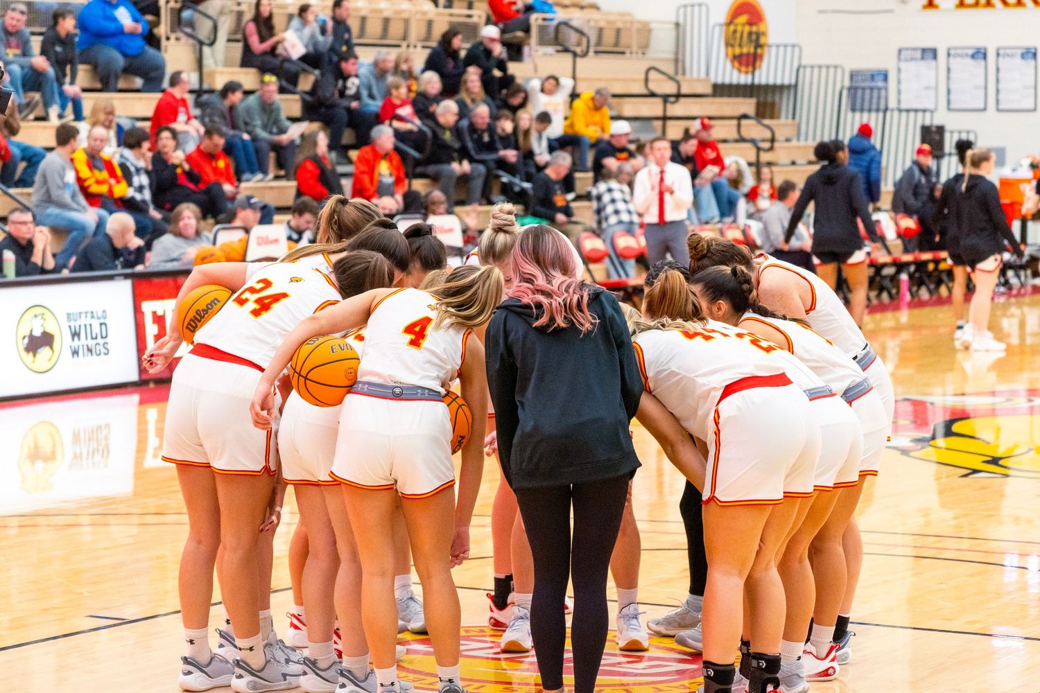 Basketball team huddles together with their coach during a timeout in a high school gymnasium.